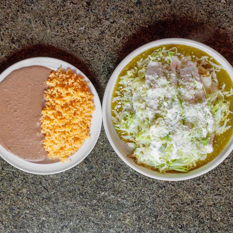 Mexican meal featuring refried beans and orange rice alongside enchiladas in green sauce topped with lettuce, cream, and cheese.