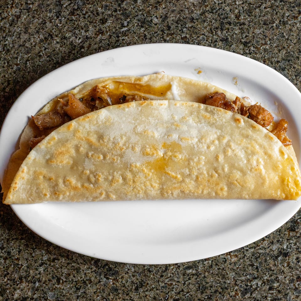 Two soft flour tacos filled with saucy browned meat on a white plate atop a speckled countertop.