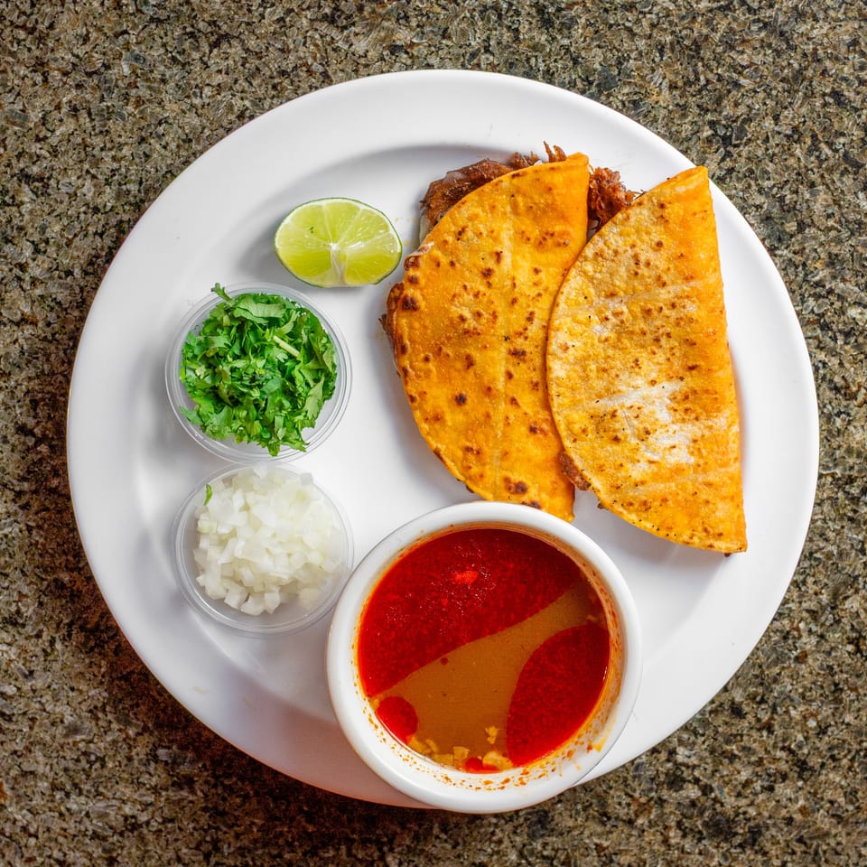 Crispy folded tacos served with lime, chopped cilantro, diced onion, and red salsa on a white plate.