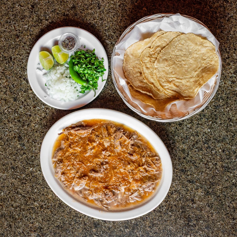 Shredded meat in savory sauce served with warm tortillas, chopped onion, cilantro, lime wedges, and green chile.