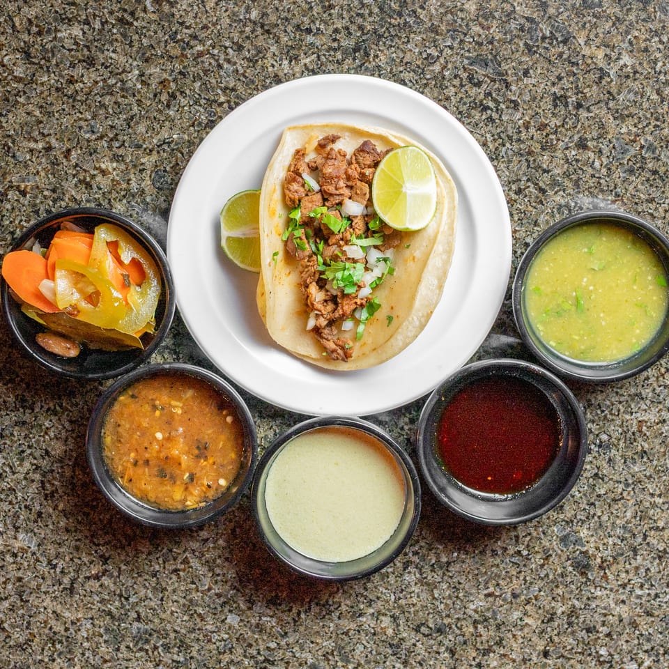 Overhead view of a Mexican taco topped with seasoned meat, onions, cilantro, and lime, surrounded by pickled vegetables and colorful salsas.