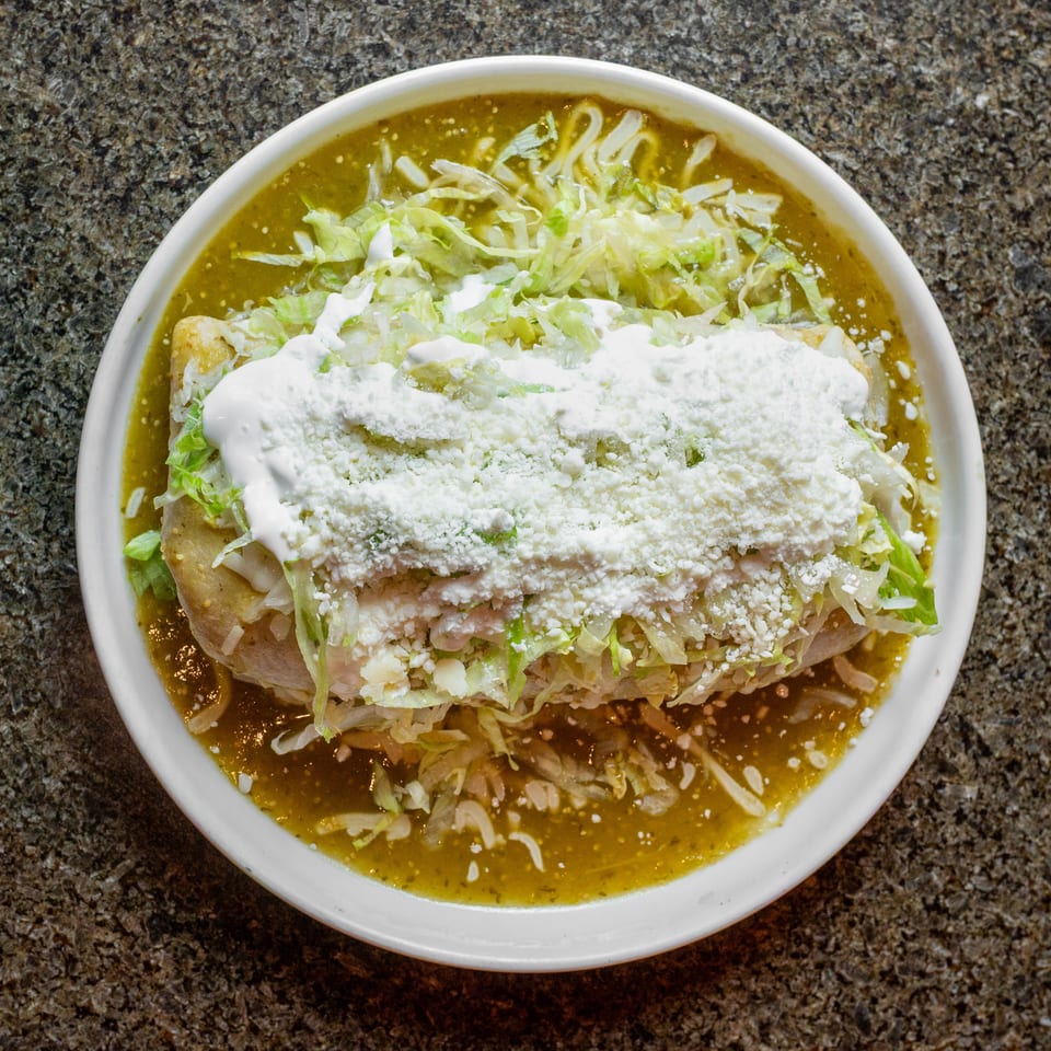 Mexican-style enchiladas topped with shredded lettuce, cream, and crumbled cheese, surrounded by green salsa on a white plate.